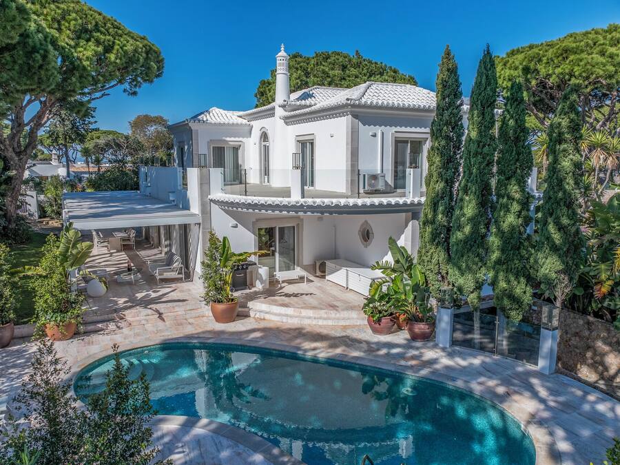 Una casa de dos pisos con piscina, rodeada de abundante vegetación y un cielo azul despejado.