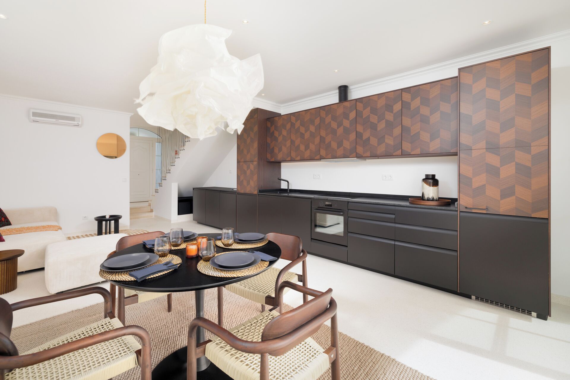 Une cuisine moderne avec un revêtement mural géométrique, un coin repas avec une table ronde et des chaises, et un escalier menant à un niveau supérieur.