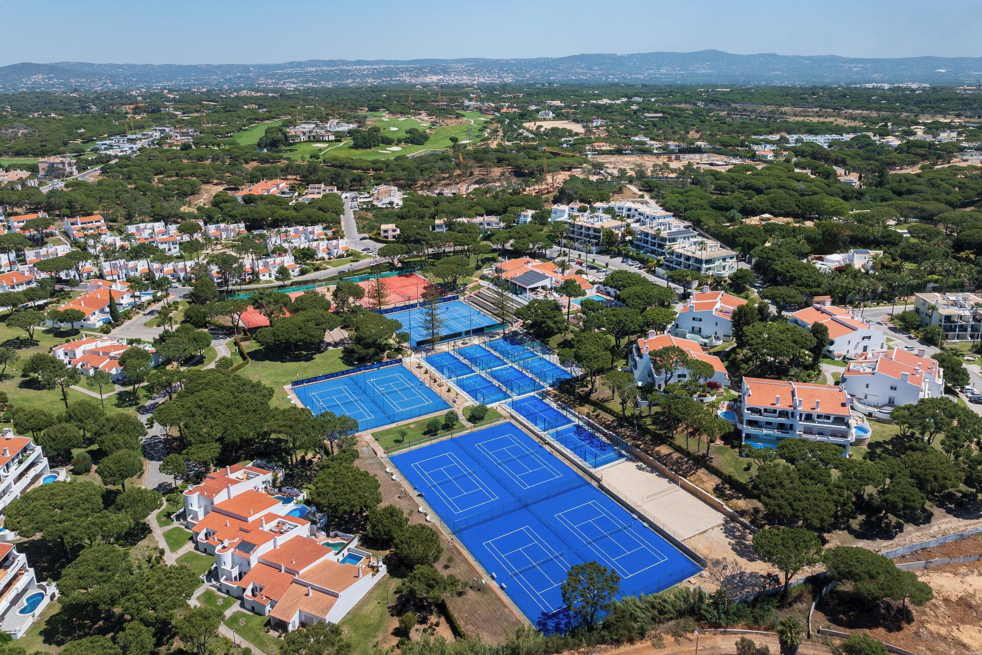 An aerial view of a residential area with multiple tennis courts, houses with red-tiled roofs, and surrounding greenery.