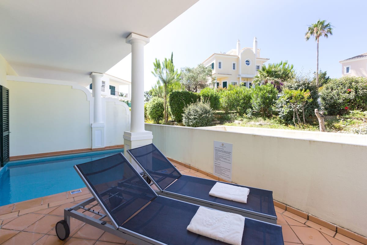 A poolside area with two sun loungers, a tiled floor, and a large building in the background.
