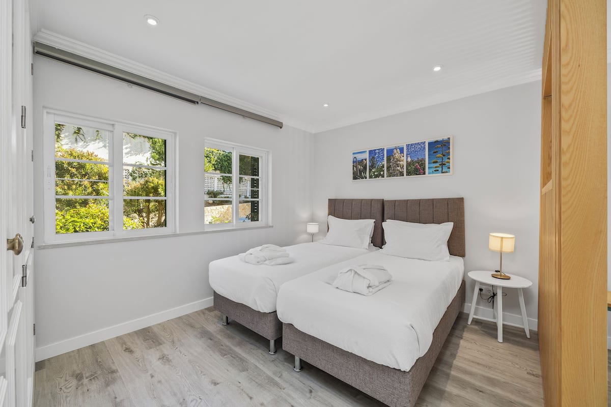 A bedroom with two beds, a window, a bedside table with a lamp, and a wall-mounted artwork.
