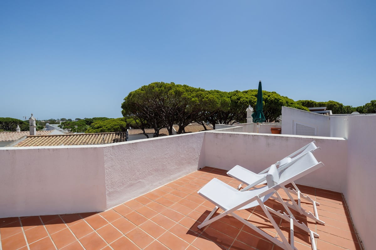 A rooftop terrace with white lounge chairs, a tiled floor, and a view of trees and buildings in the background.