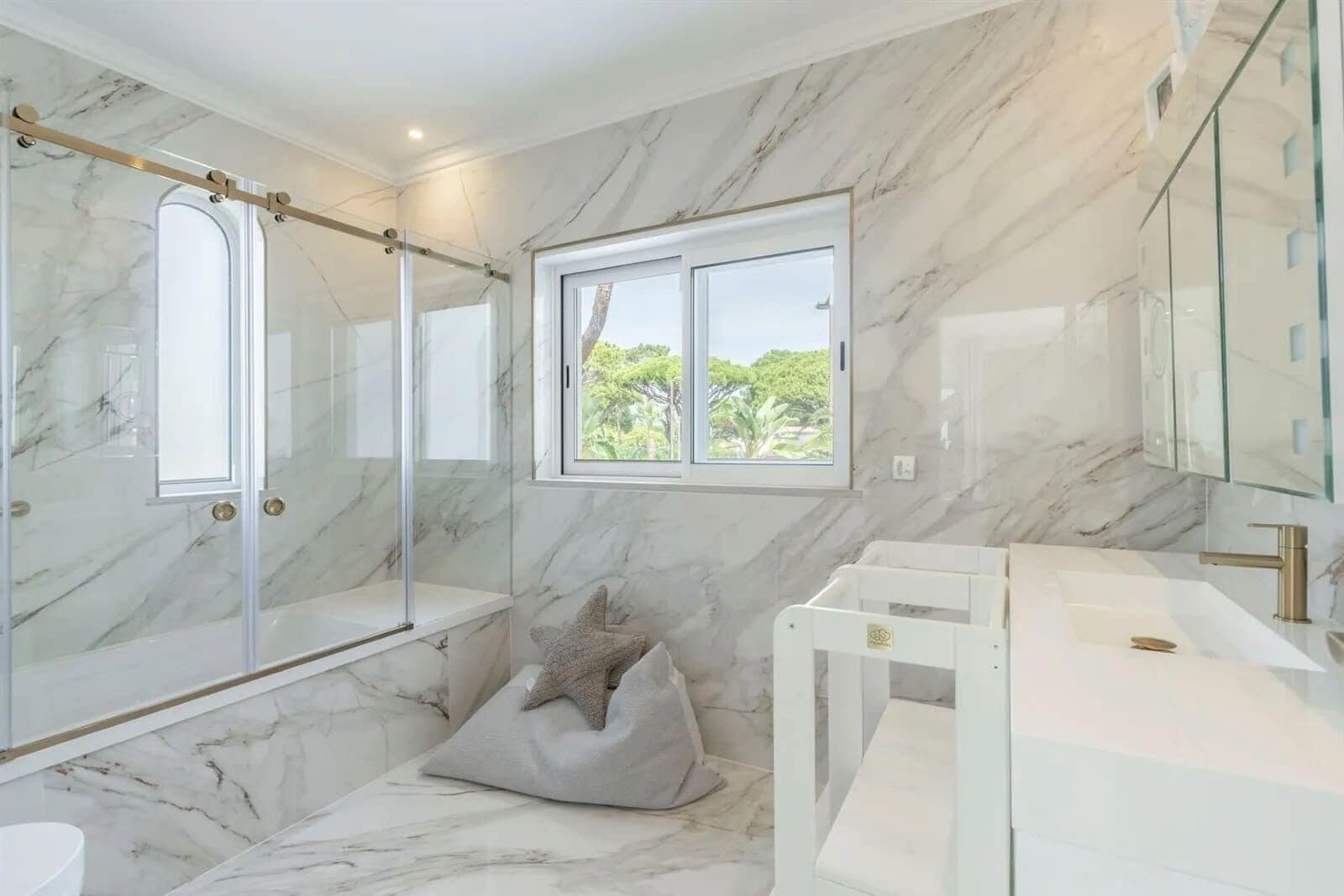 A bathroom with marble walls, a glass shower enclosure, a window with a view, and a white sink with a gold faucet.