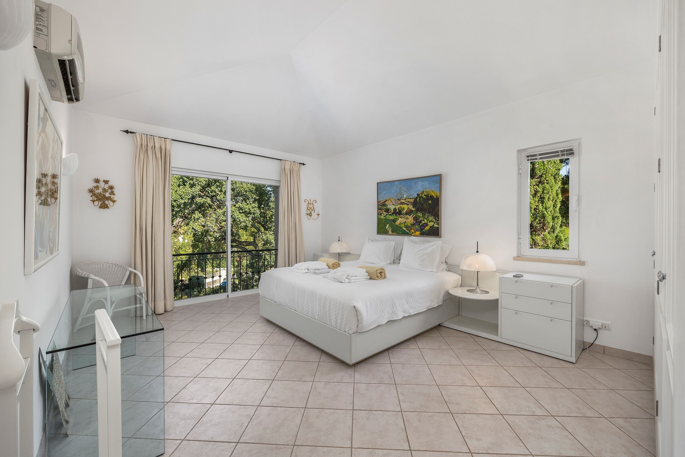 Un dormitorio con cama, mesitas de noche, una mesa de vidrio, una silla, un balcón con vistas y un cuadro en la pared.