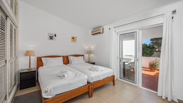 Una cama doble con sábanas blancas y dos mesitas de noche, una puerta corrediza al balcón y una pequeña planta en maceta en el balcón.
