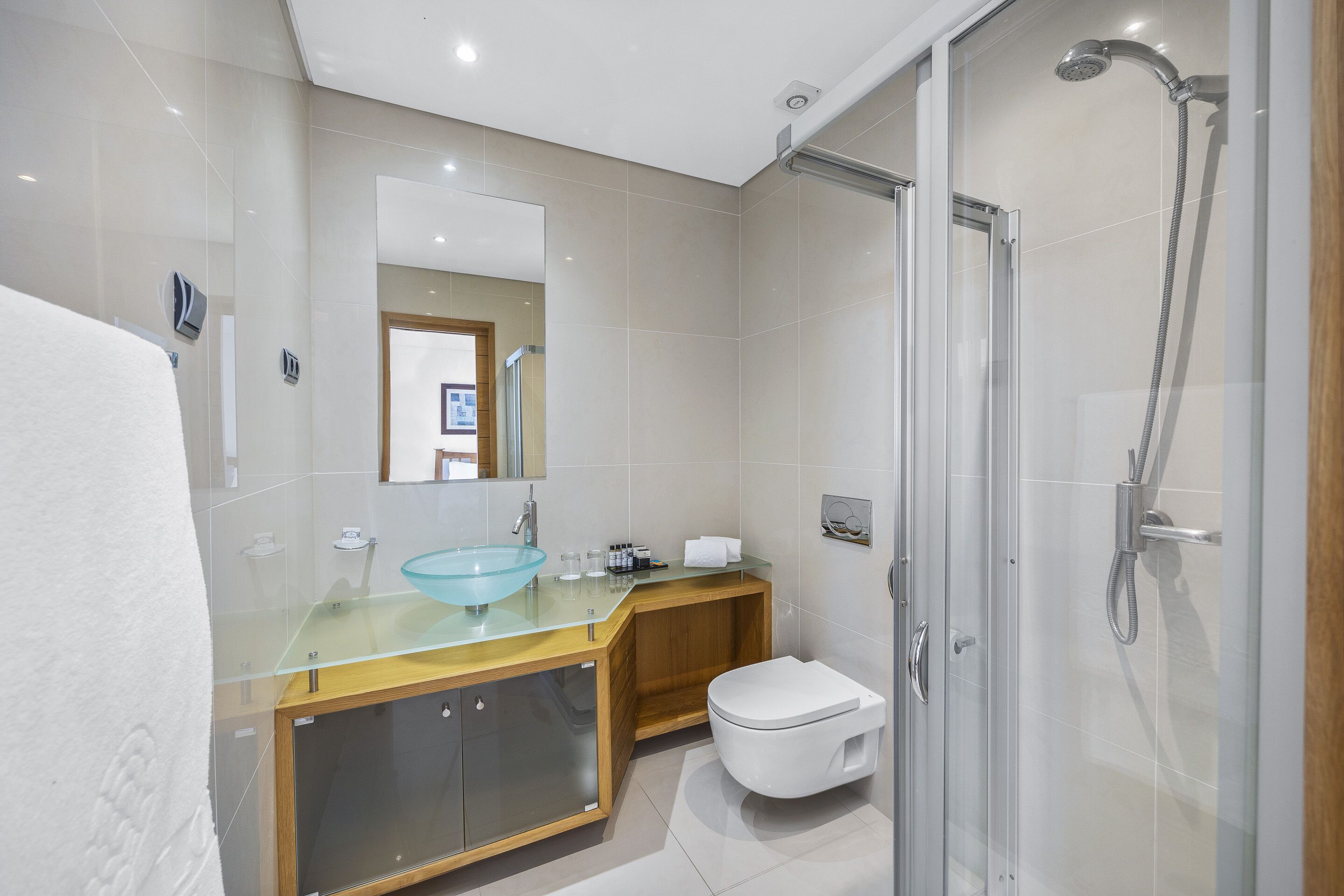 Un baño moderno con lavamanos de vidrio, tocador de madera, inodoro blanco y zona de ducha con cerramiento de vidrio.