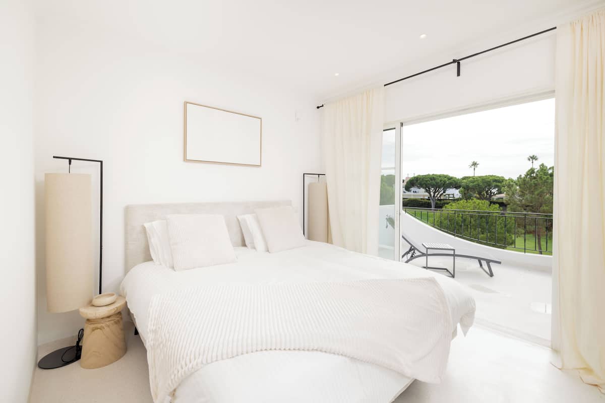 A modern bedroom with a large bed, a bedside table, a lamp, and a view of the outdoors.