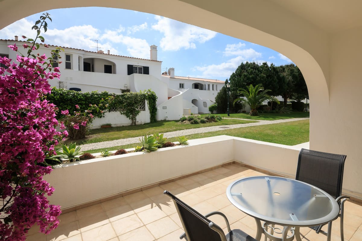 A patio with a table and chairs, a view of a white building with a garden, and a clear sky.