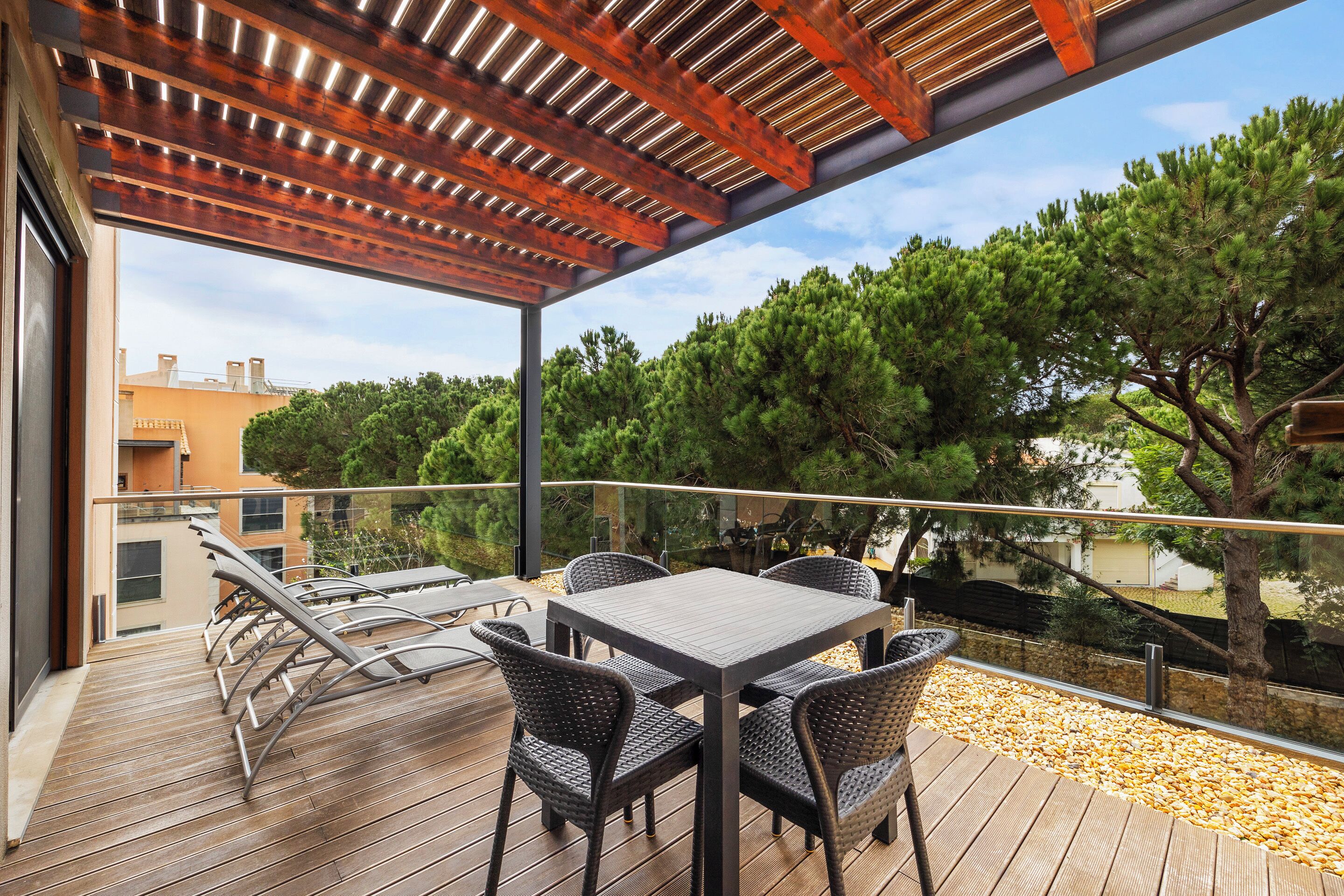A wooden deck with a table and chairs, overlooking a garden with trees and a building in the background.