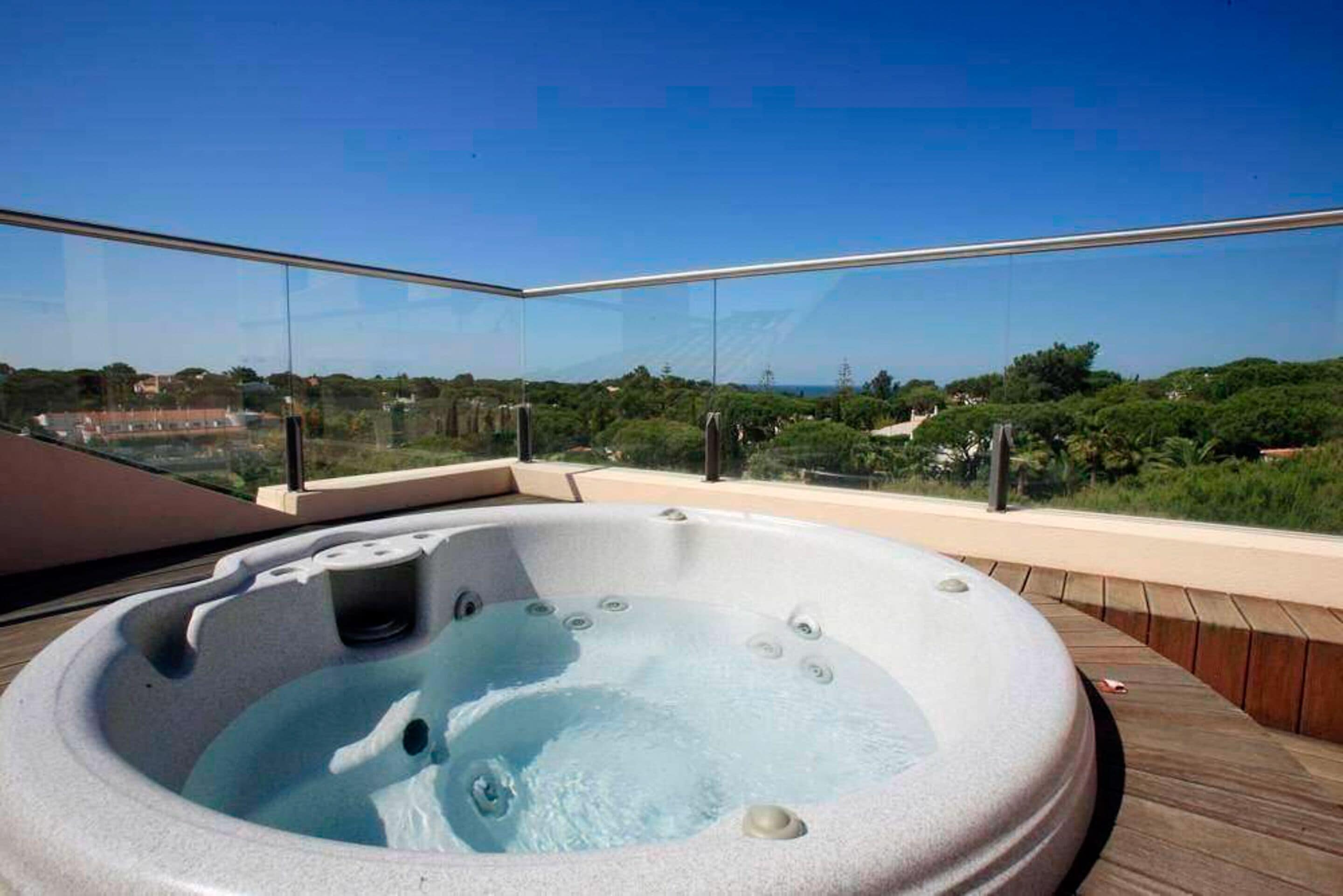 A hot tub on a balcony with glass railings, overlooking a landscape with trees and buildings.