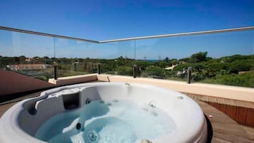 A hot tub on a balcony with glass railings, overlooking a landscape with trees and buildings.