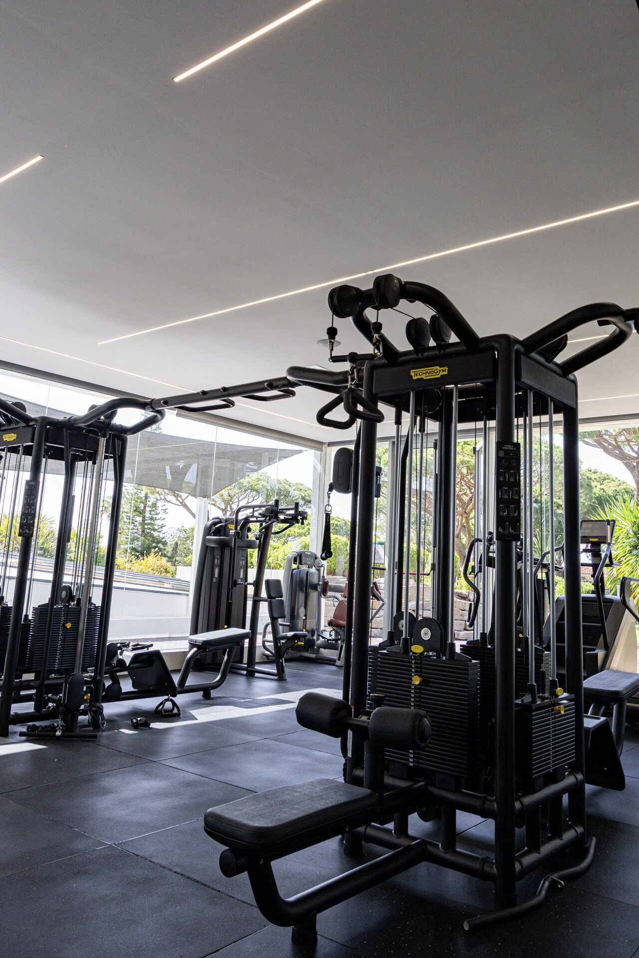 Un gimnasio con diversas máquinas de ejercicio, bancos y una zona cubierta con ventanas amplias.