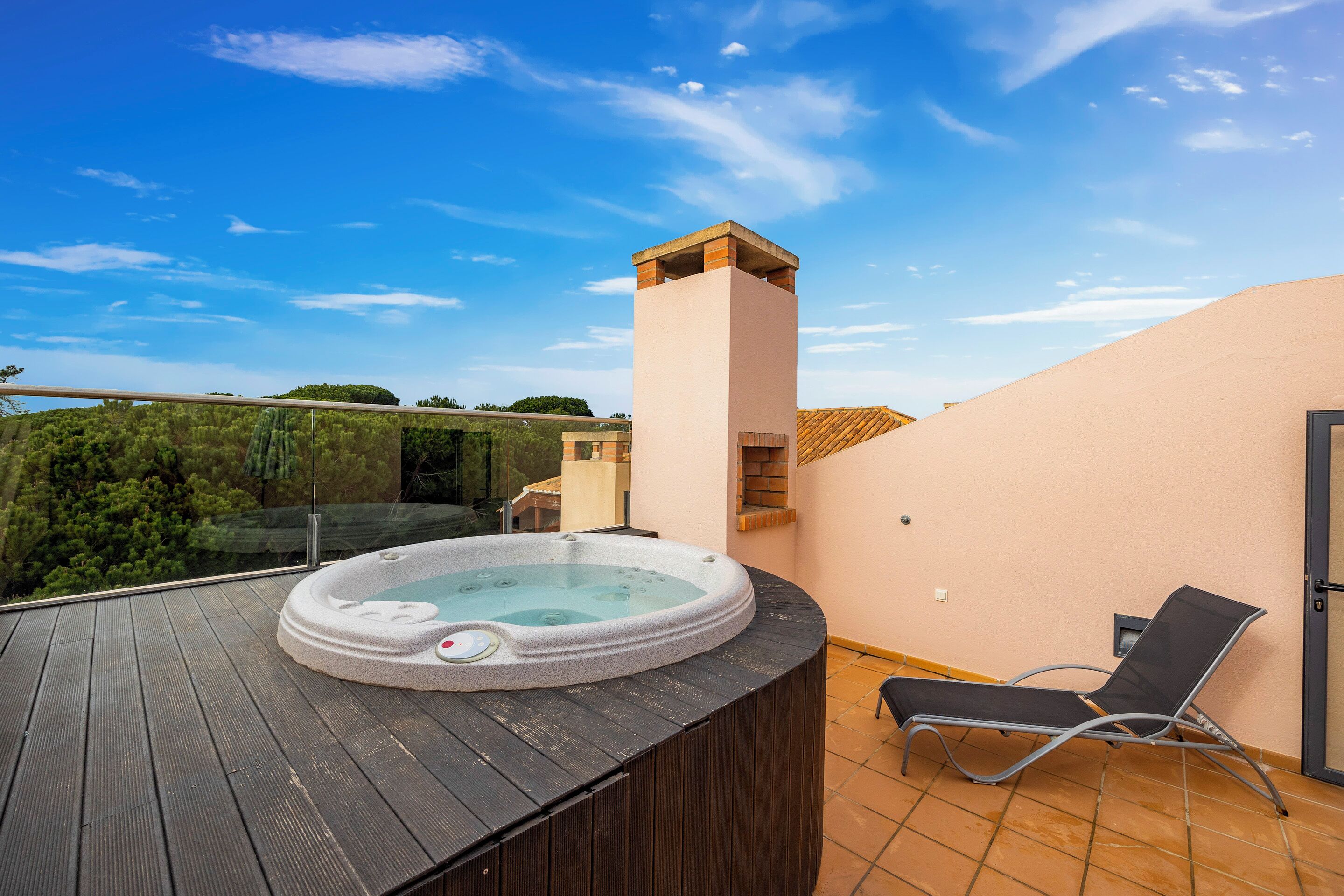 A hot tub on a wooden deck with a chair, a tall chimney, and a view of trees and a building in the background.