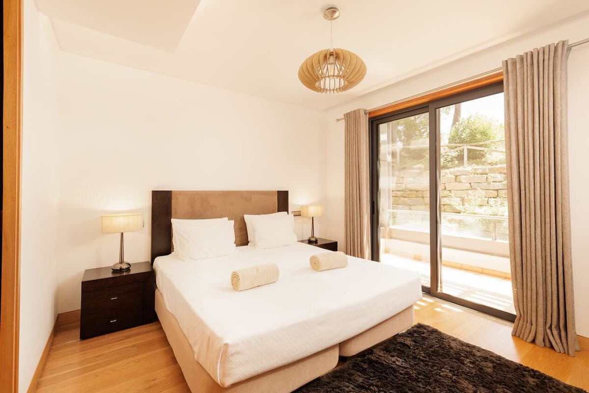 A bedroom with a large bed, two bedside tables with lamps, a dark rug, and a view through a glass door.