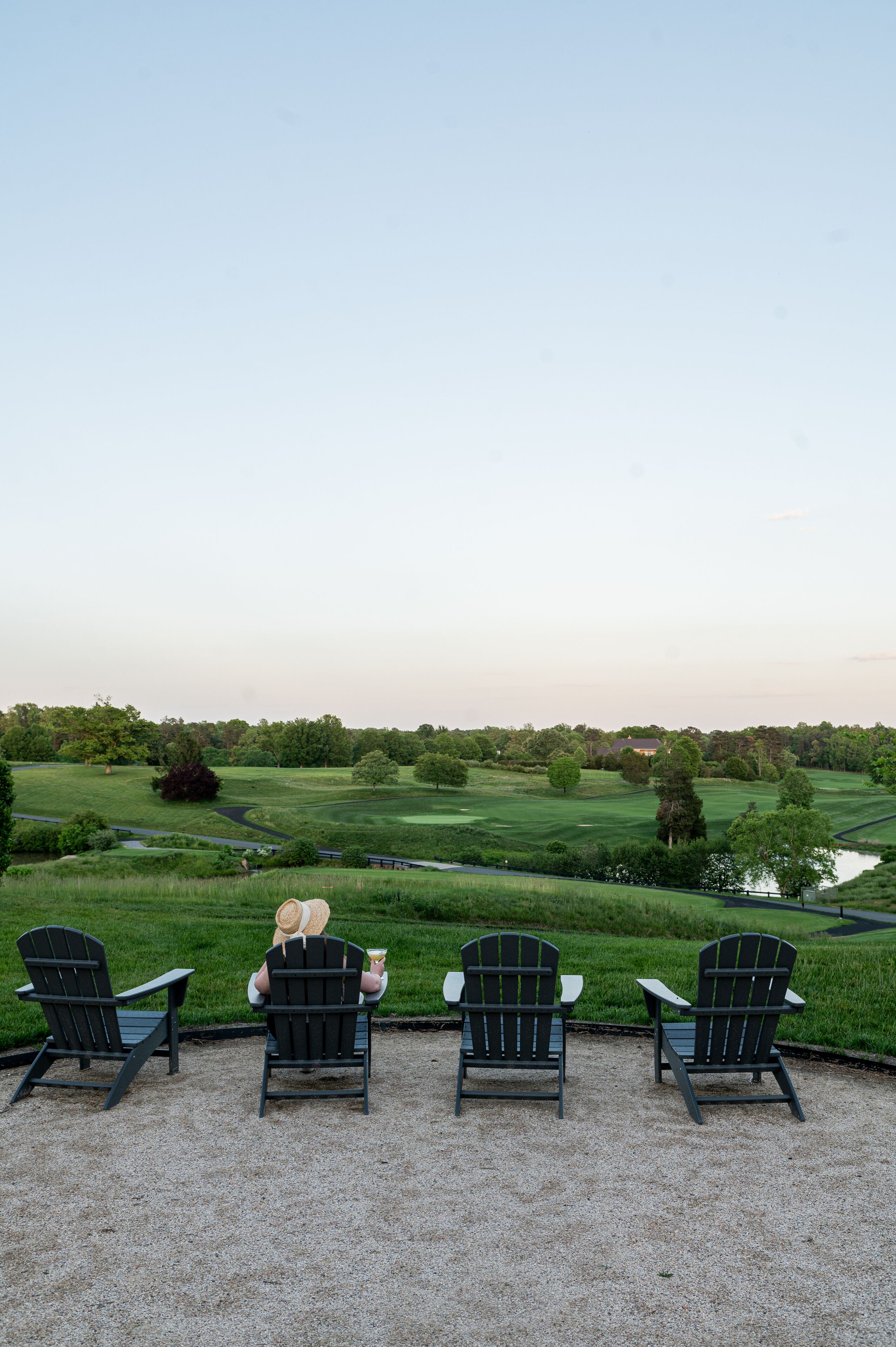 Four Adirondack chairs facing a golf course with a pond and trees in the background.