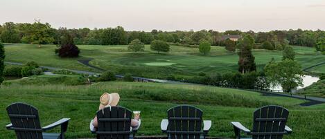 Four Adirondack chairs facing a golf course with a pond and trees in the background.