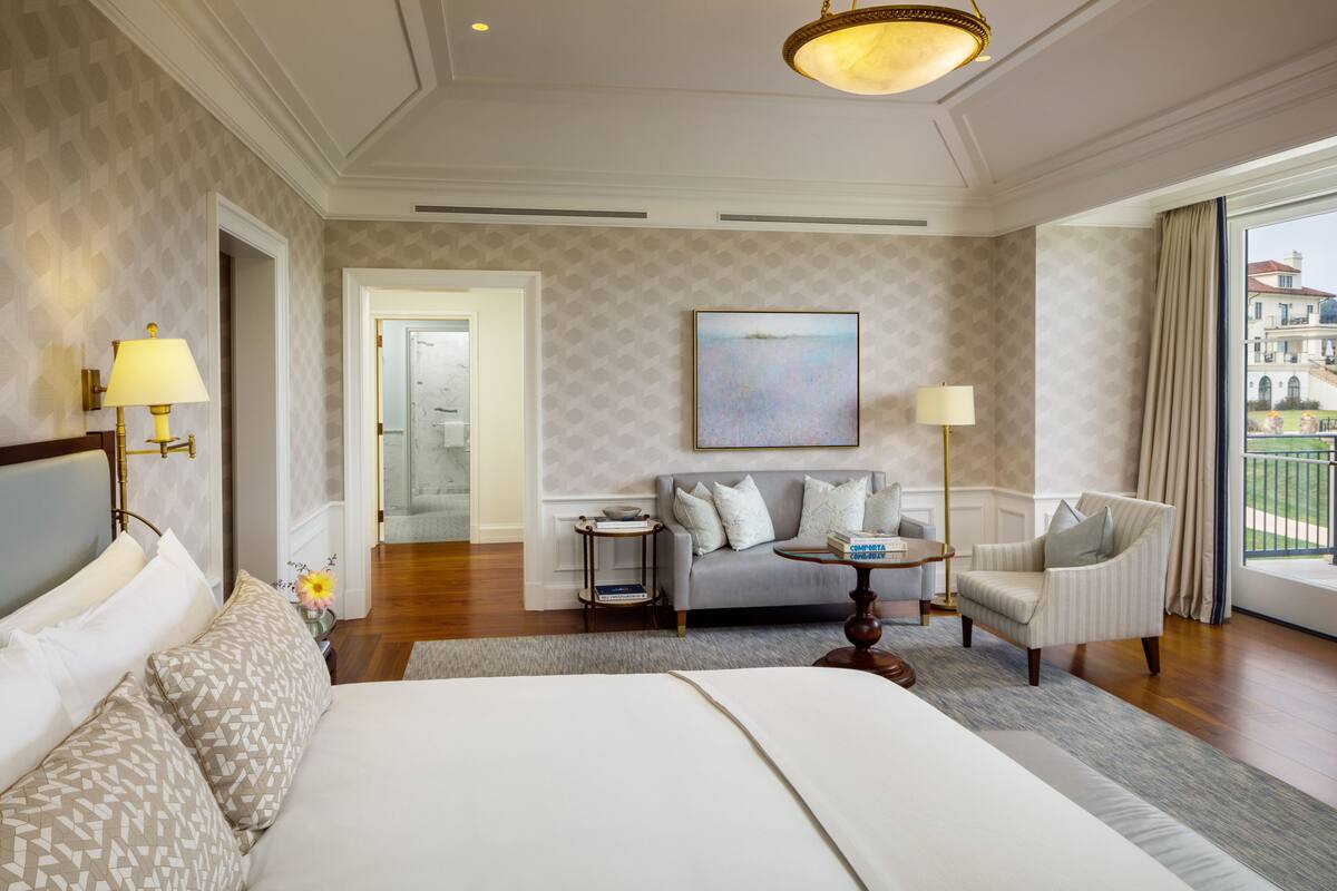 A hotel room with a large bed, a sofa, a chair, a small table, and a painting on the wall.