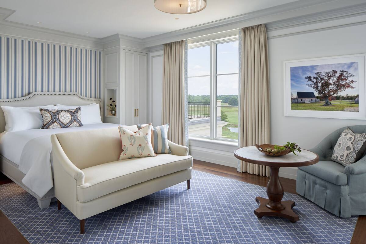 A hotel room with a large bed, a sofa, a round table with a bowl, and a chair.