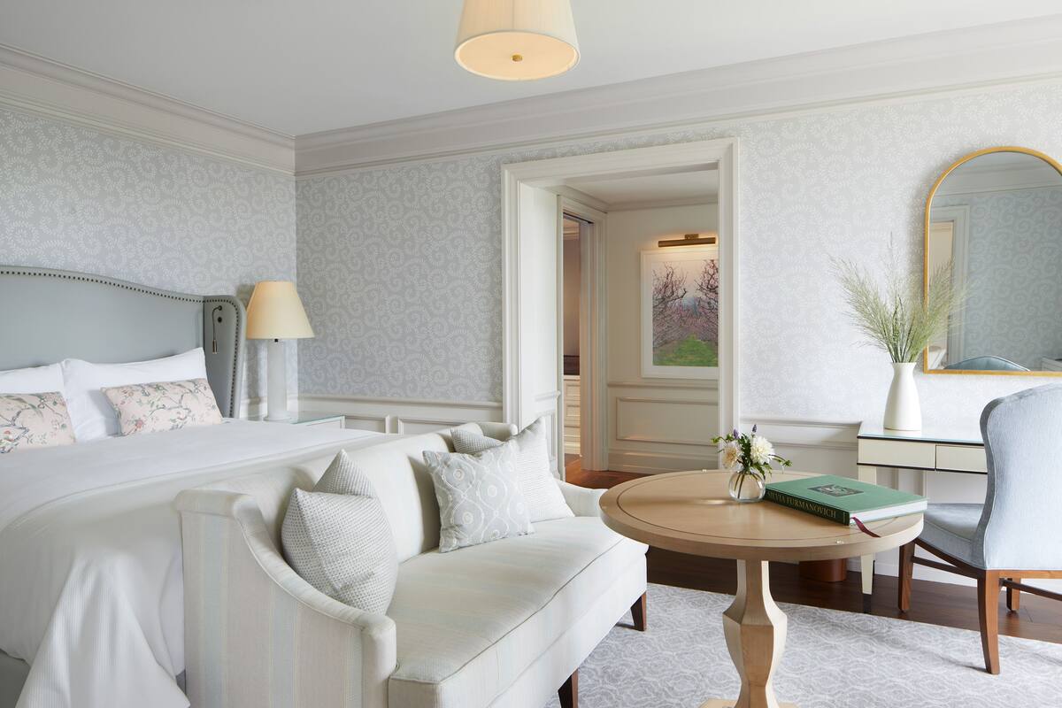 A hotel room with a large bed, a sofa, a round table with a book and vase, and a mirror.