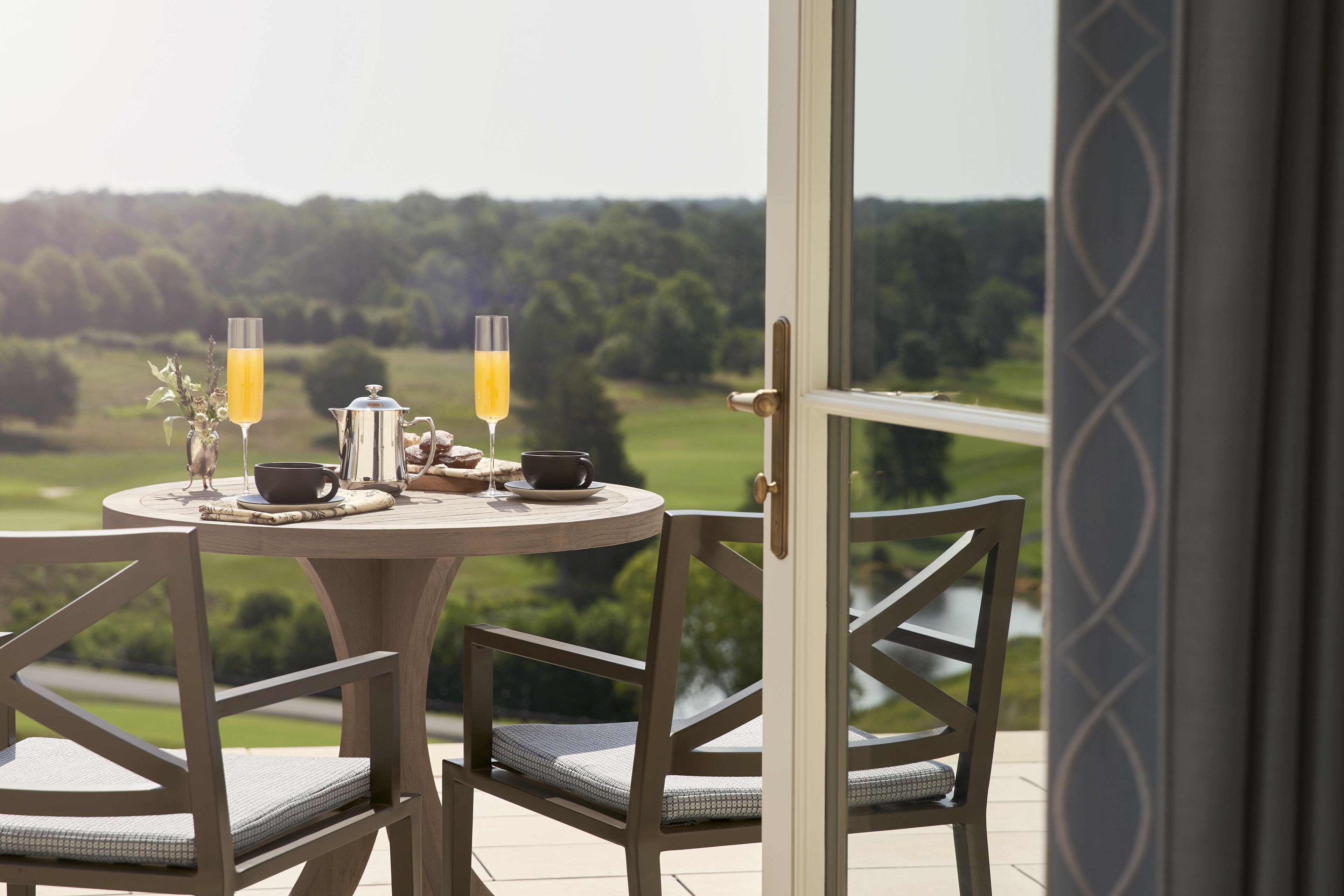A dining table with two glasses of orange juice, a teapot, and a cup, set on a balcony overlooking a green landscape.
