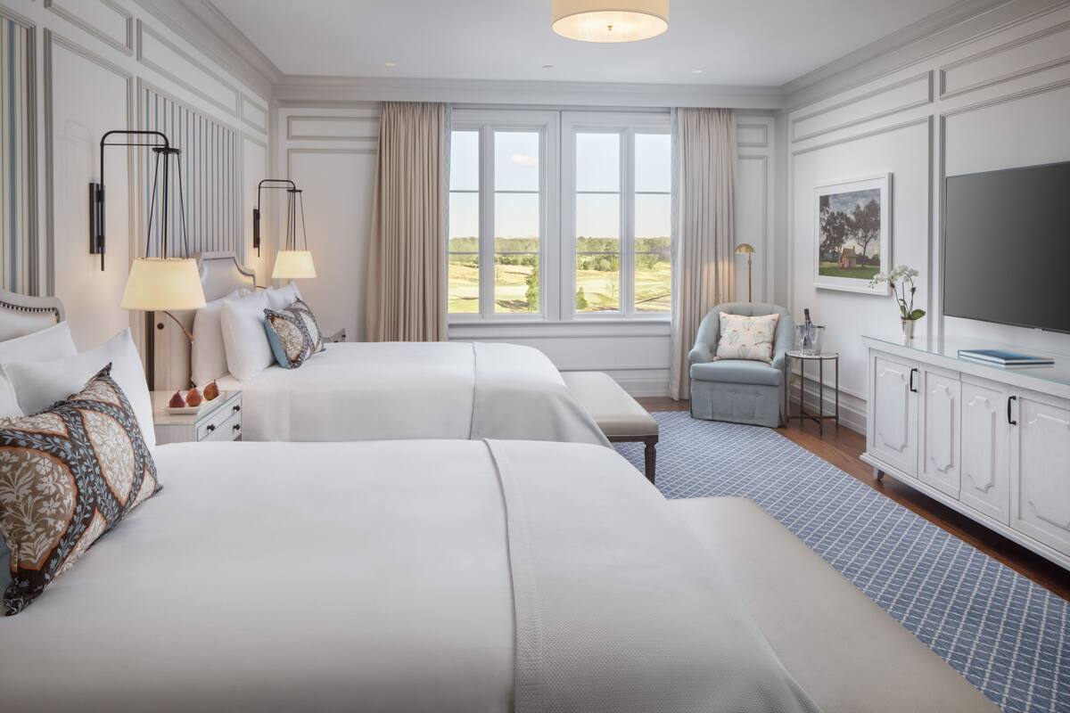 A hotel room with two beds, a TV, a desk with a lamp, and a view of the outdoors.