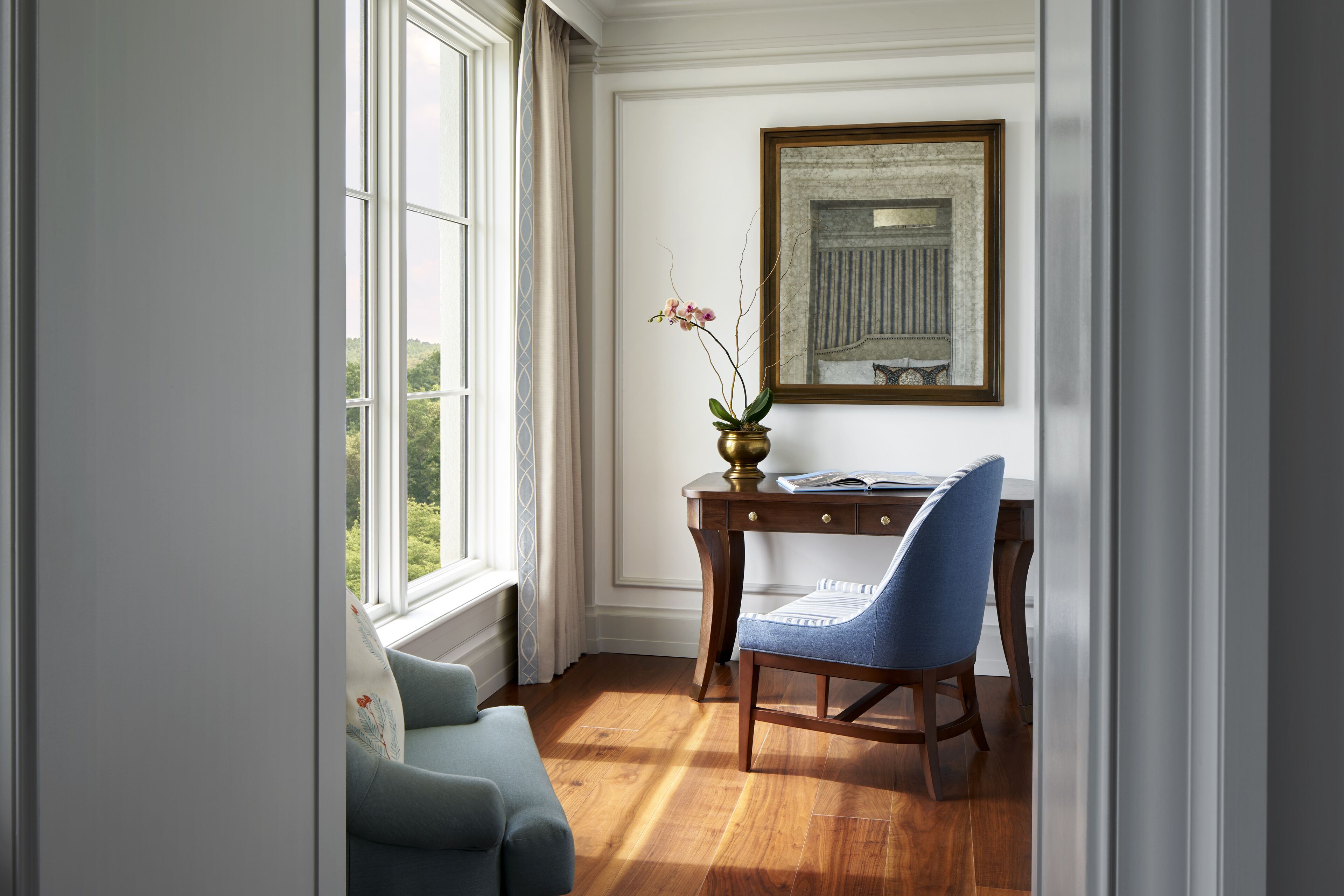 Una habitación con un escritorio de madera, una silla azul, un florero con flores y un cuadro en la pared.