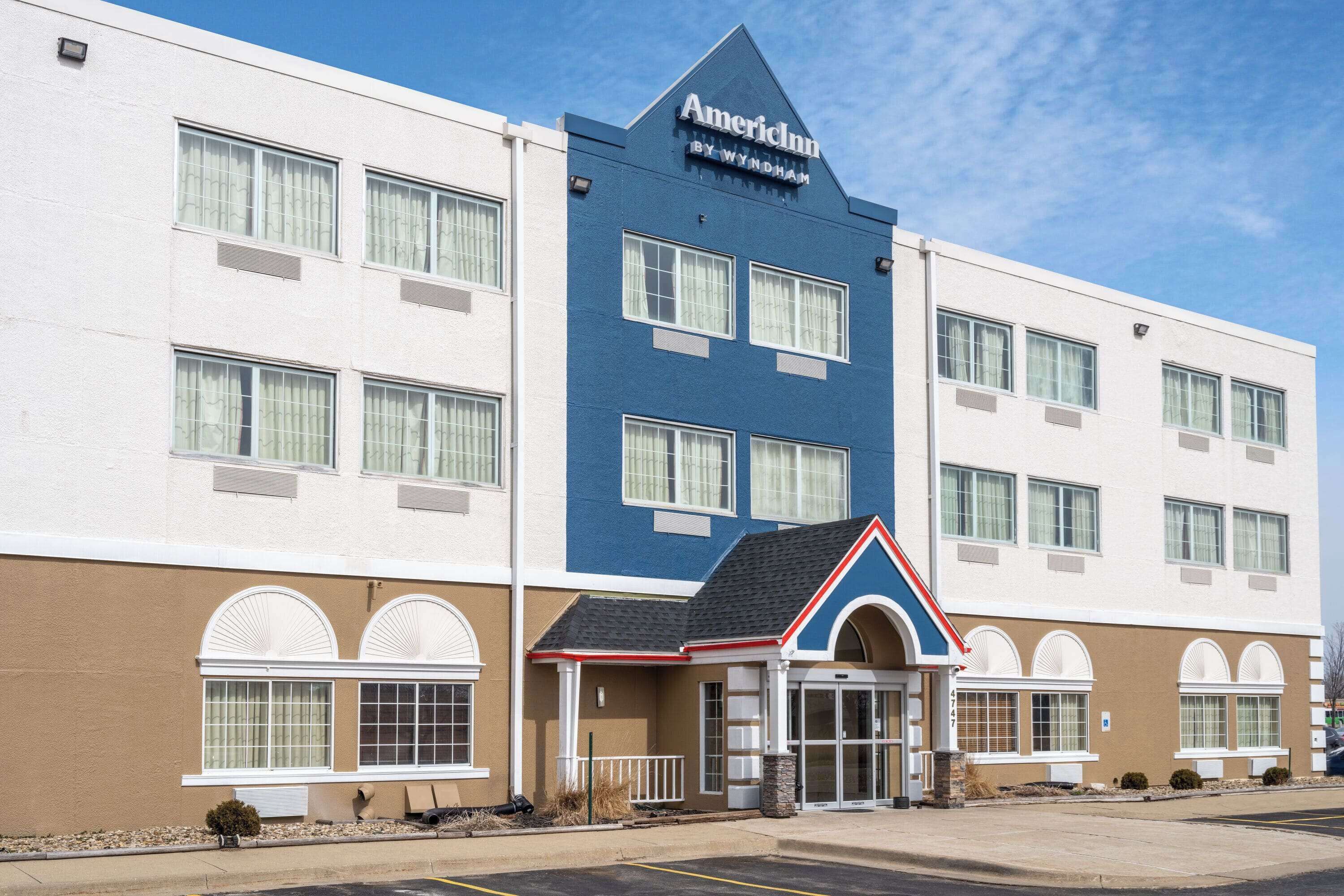 Un hotel de varios pisos con fachada azul y blanca, un llamativo letrero de American Inn y una entrada central con toldo rojo y blanco.
