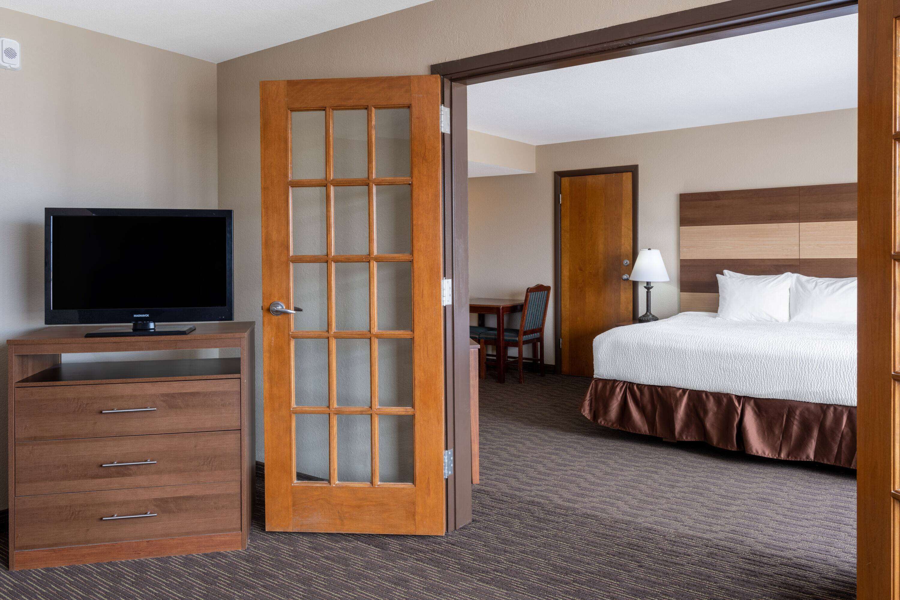 A hotel room with a wooden dresser, a television, a bed with white bedding, and a wooden-framed door.