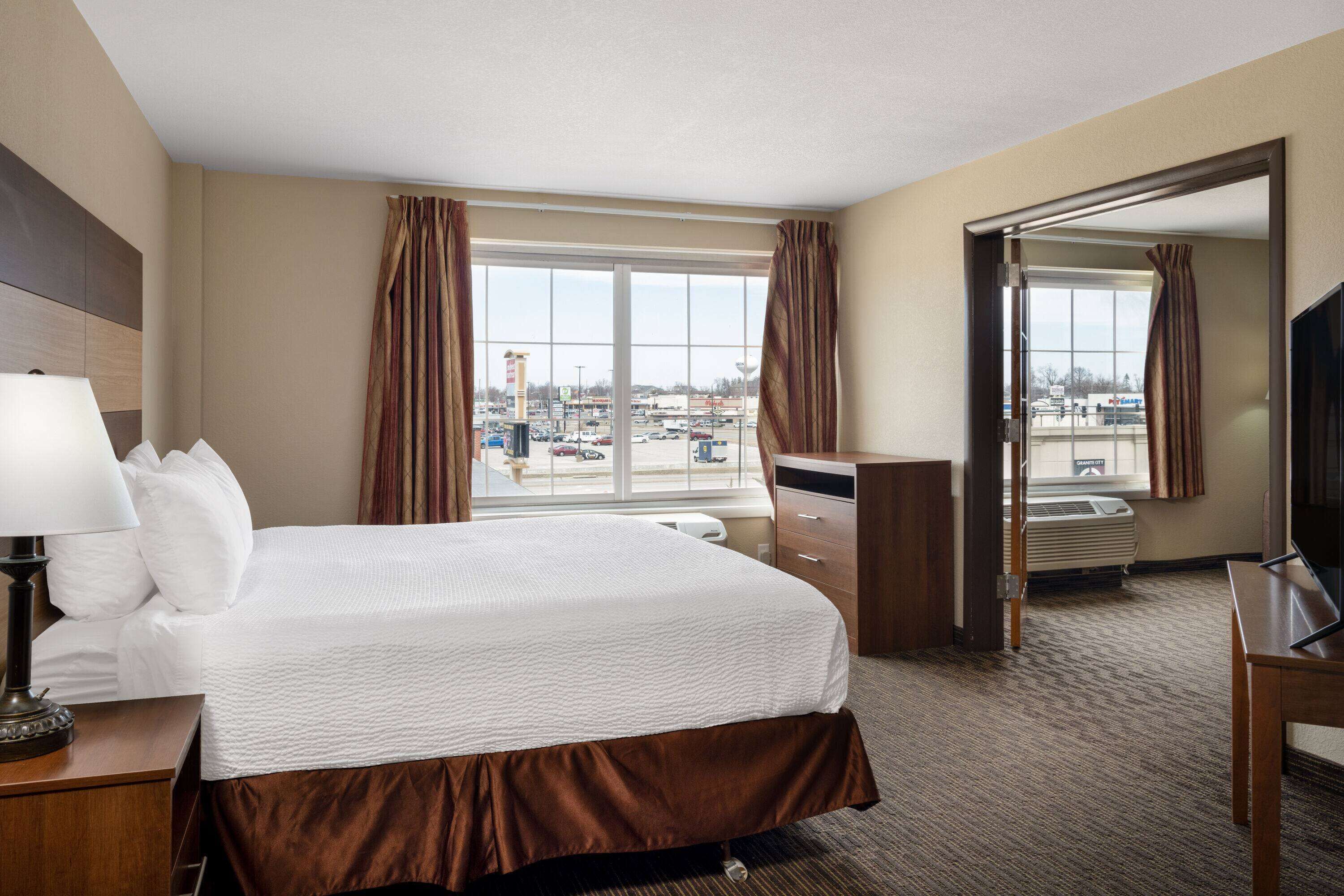 A hotel room with a large bed, a desk with a lamp, a TV, and a view of a parking lot through the window.