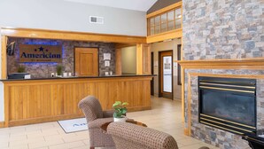 A reception area with a wooden counter, a stone wall, and a fireplace. There are two patterned chairs and a small table with a plant. - AmericInn by Wyndham Cedar Rapids North (Cedar Rapids)