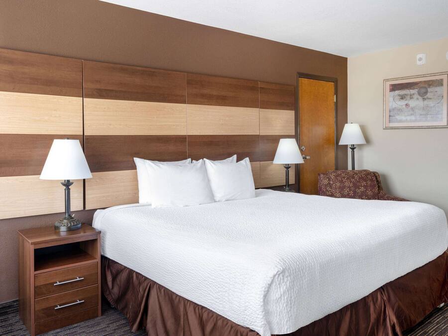 A hotel room with a large bed, two bedside tables with lamps, a wooden headboard, a brown chair, and a framed picture on the wall.