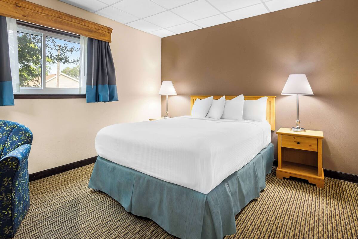 A hotel room with a large bed, two bedside tables with lamps, a chair, and a window with curtains.