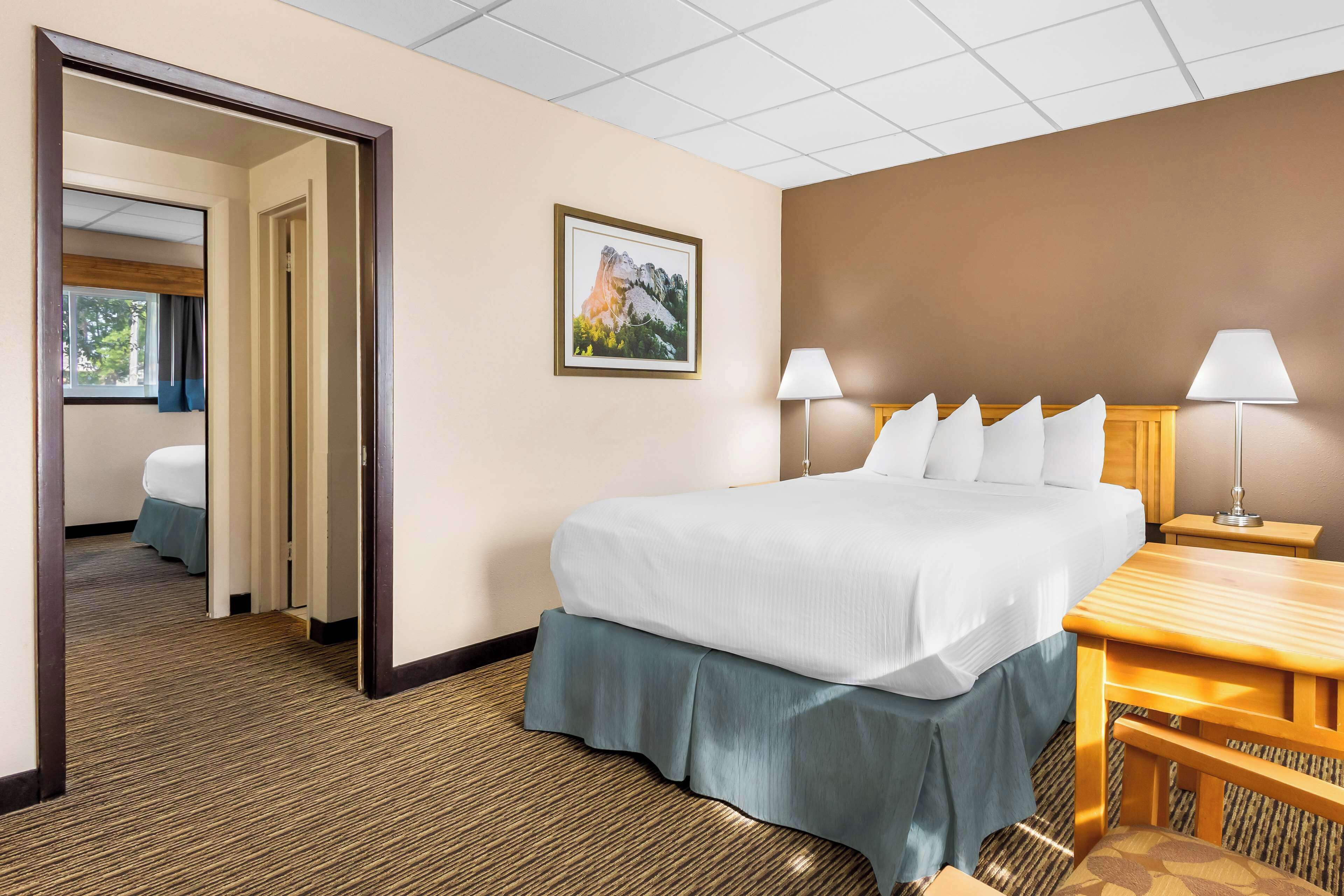 A hotel room with a large bed, a desk, two lamps, a framed picture on the wall, and a carpeted floor.