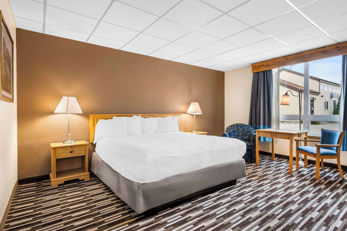 A hotel room with a large bed, two bedside tables with lamps, a desk with a chair, and a view of the outside through a large window.