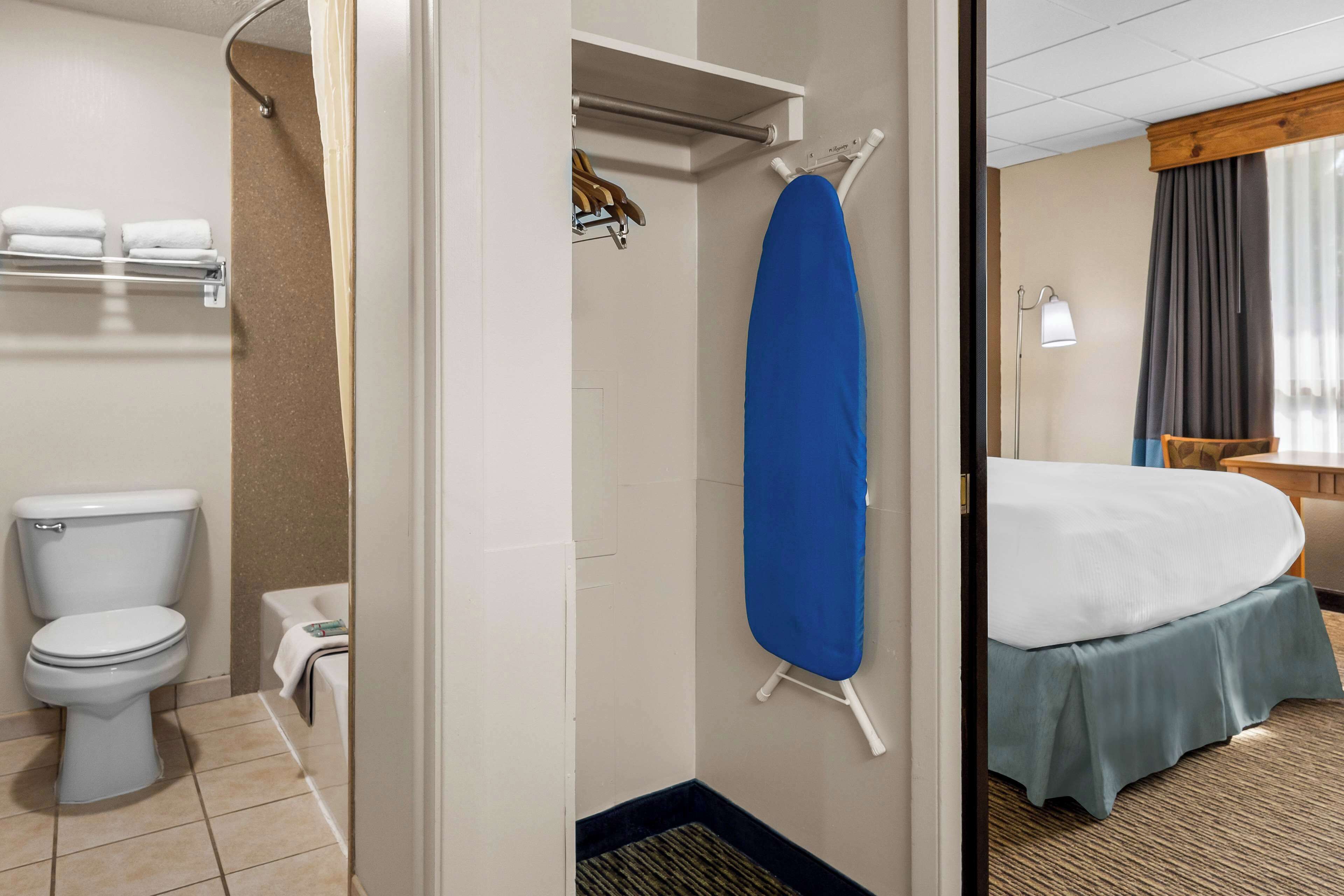 A hotel bathroom with a toilet, a shower, and a bed with a blue bed skirt.