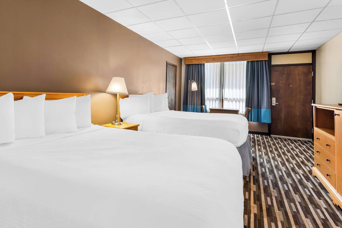A hotel room with two beds, a wooden headboard, a nightstand with a lamp, a dresser, and a window with curtains.