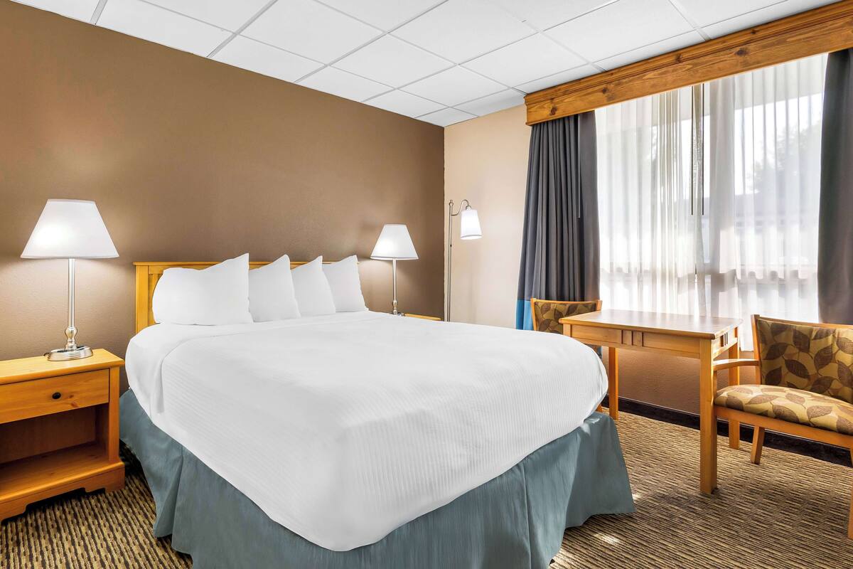 A hotel room with a large bed, two bedside tables with lamps, a wooden desk, a chair, and a large window with curtains.