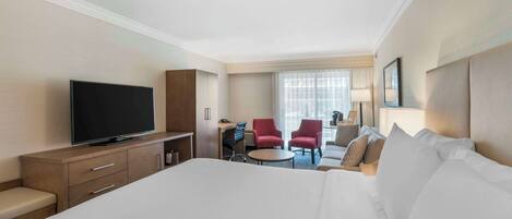 A hotel room with a large bed, a television, a desk with a chair, and two red armchairs.