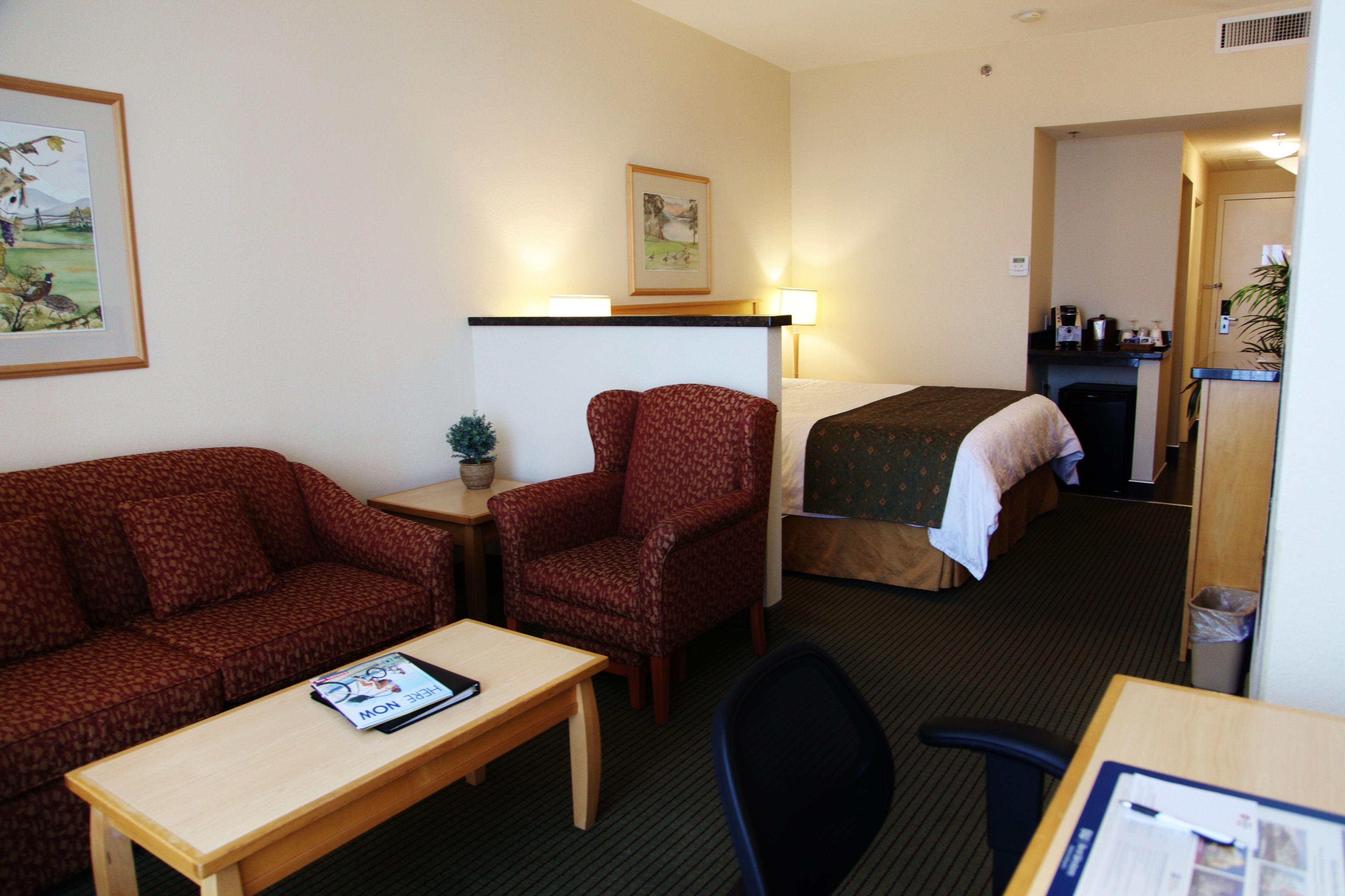 Ein Hotelzimmer mit Bett, einer Couch, einem kleinen Tisch, einem Sessel, einem Schreibtisch, einer Lampe, einem Papierkorb und einem gerahmten Bild an der Wand.