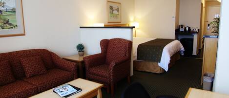 Ein Hotelzimmer mit Bett, einer Couch, einem kleinen Tisch, einem Sessel, einem Schreibtisch, einer Lampe, einem Papierkorb und einem gerahmten Bild an der Wand.
