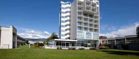 A modern multi-story hotel with a central glass facade, surrounded by a well-maintained lawn and a clear blue sky.