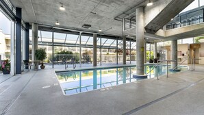 A large indoor swimming pool with clear glass walls, concrete pillars, and a spacious area with seating and plants.