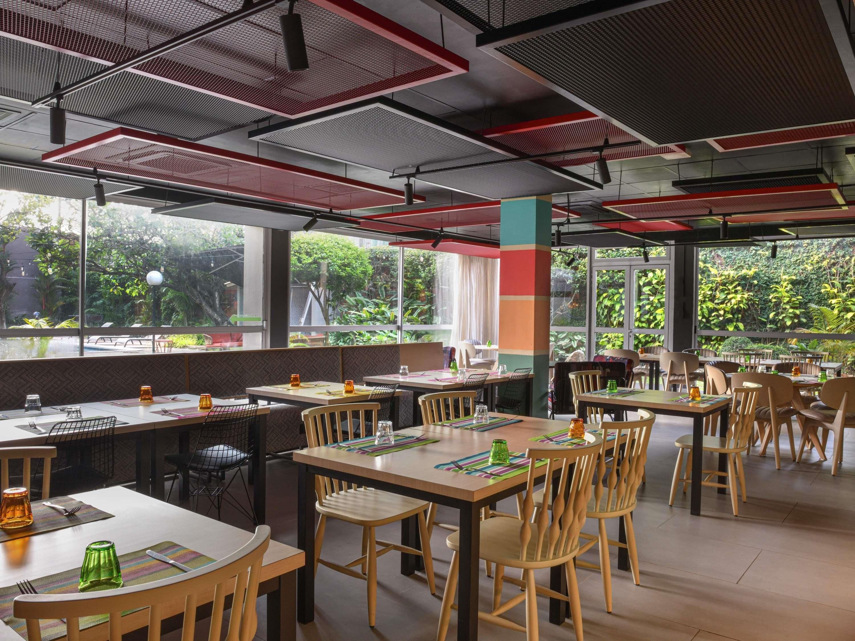 A modern restaurant with high ceilings, large windows, and a view of greenery outside. Tables are arranged with chairs, and the interior has a contemporary design.