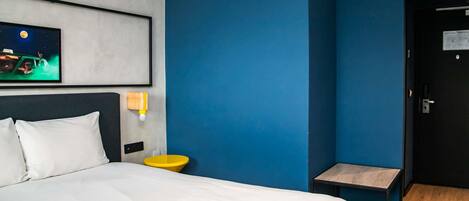 A hotel room with a bed, a nightstand, a small table, a wall-mounted light, and a door.