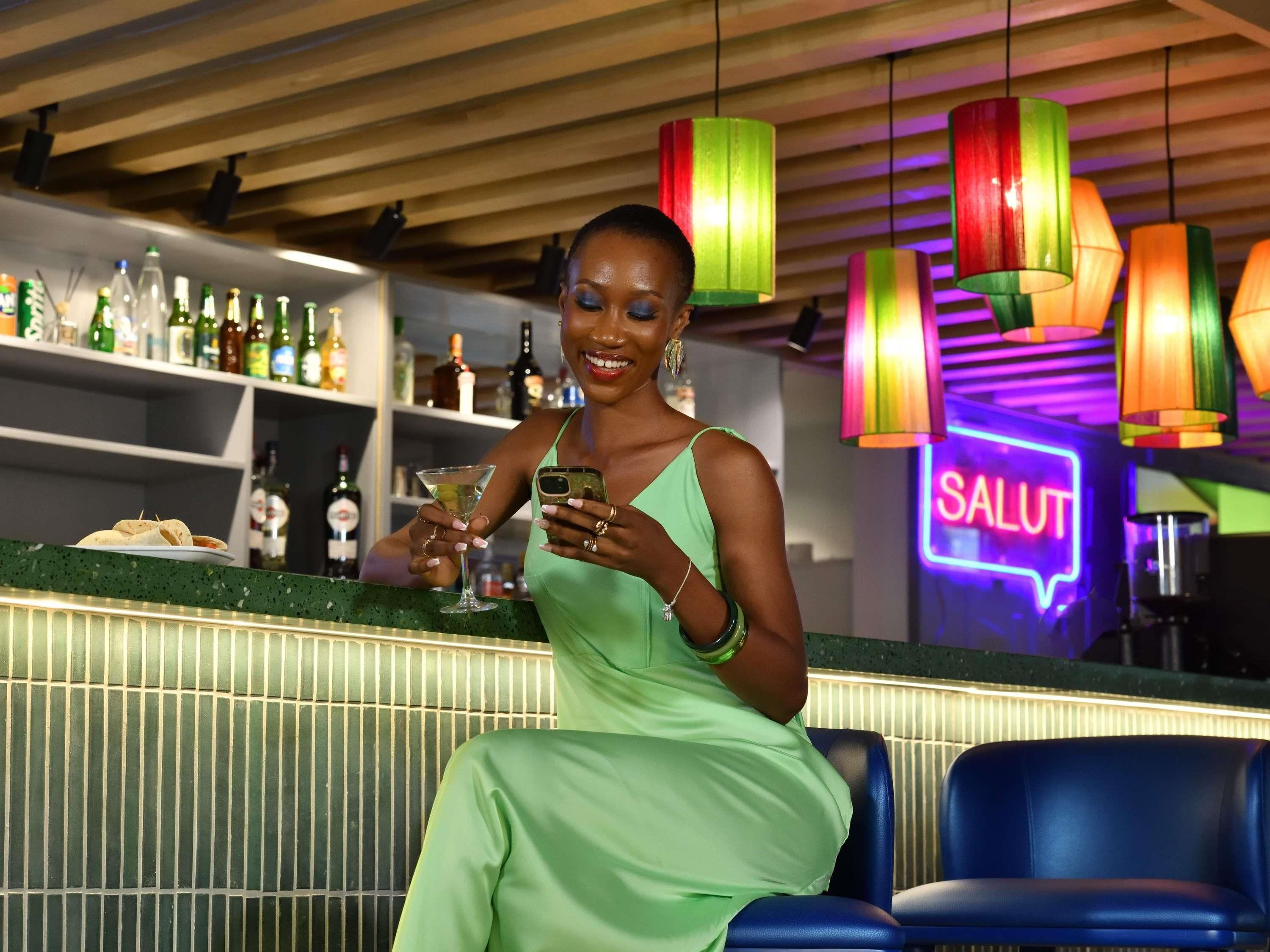 A person seated at a bar with a neon sign reading "SALUT" in the background.