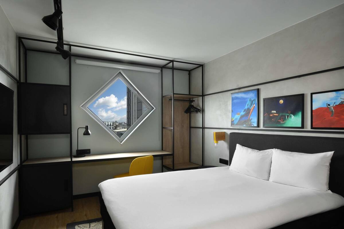 A modern hotel room with a bed, a desk, and a large window.