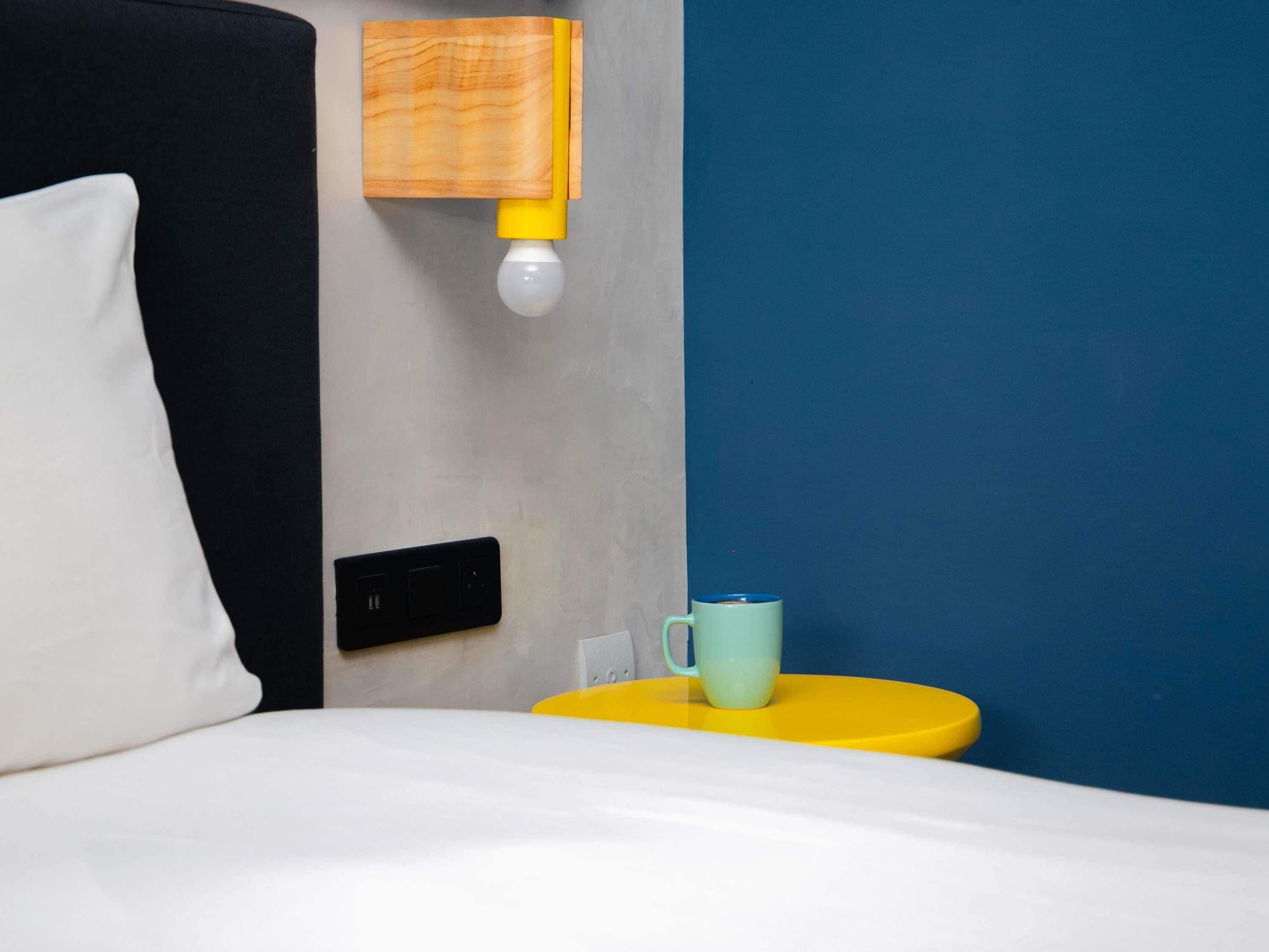 A bed with a white pillow, a yellow bedside table with a light blue mug, and a wall-mounted light fixture.