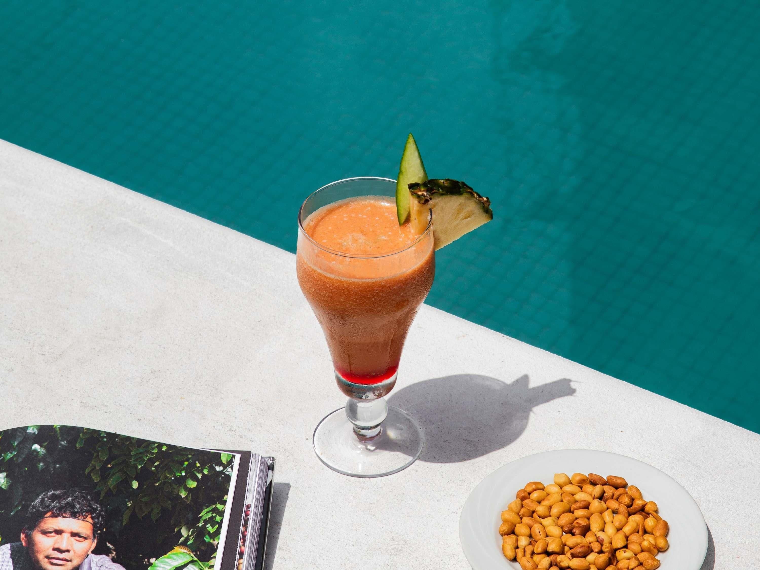 A glass of drink with a garnish, a plate of peanuts, and a CD on a table by a pool.