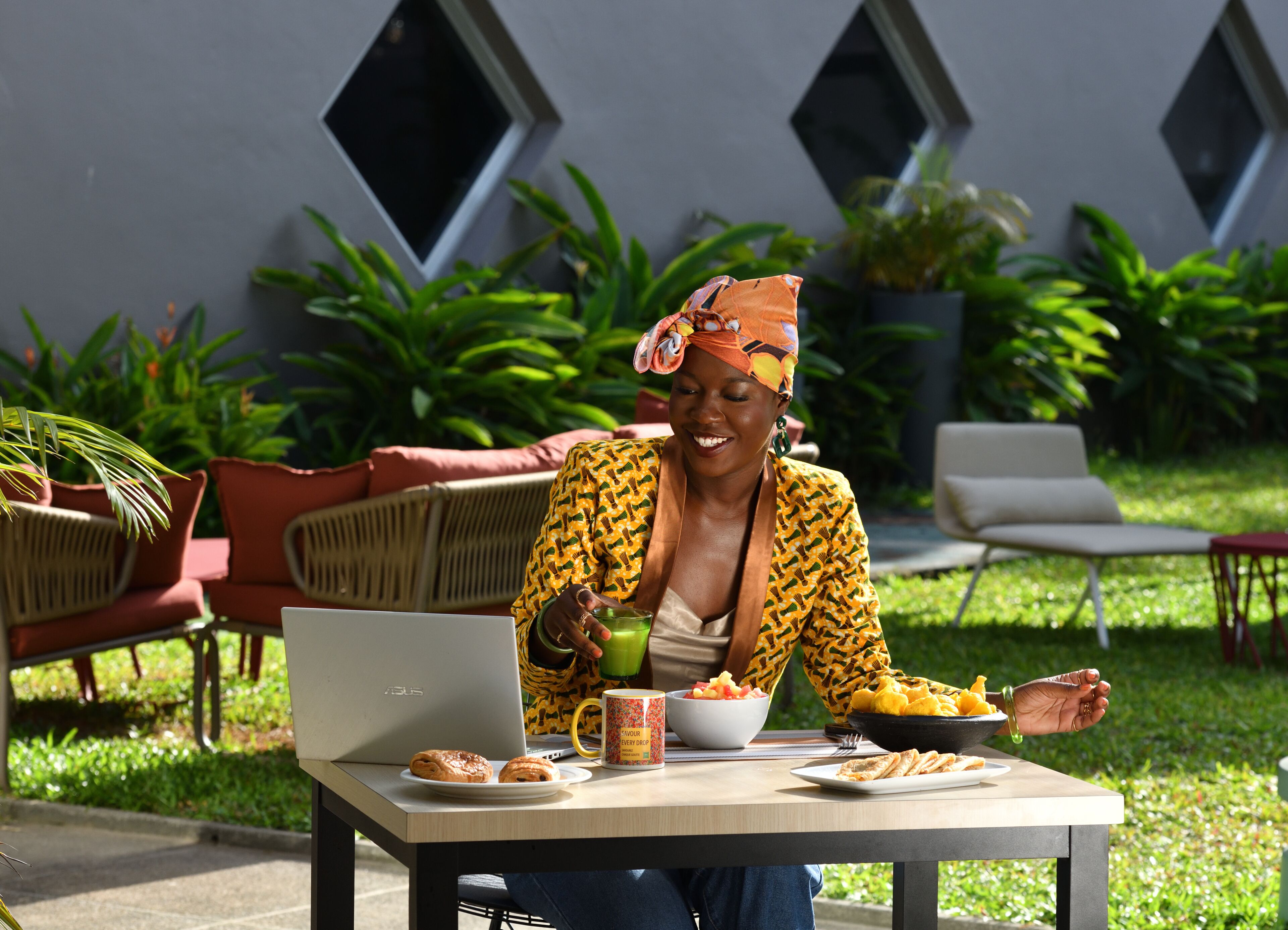 Una persona sentada en una mesa al aire libre, usando una computadora portátil, un plato de comida y una bebida.