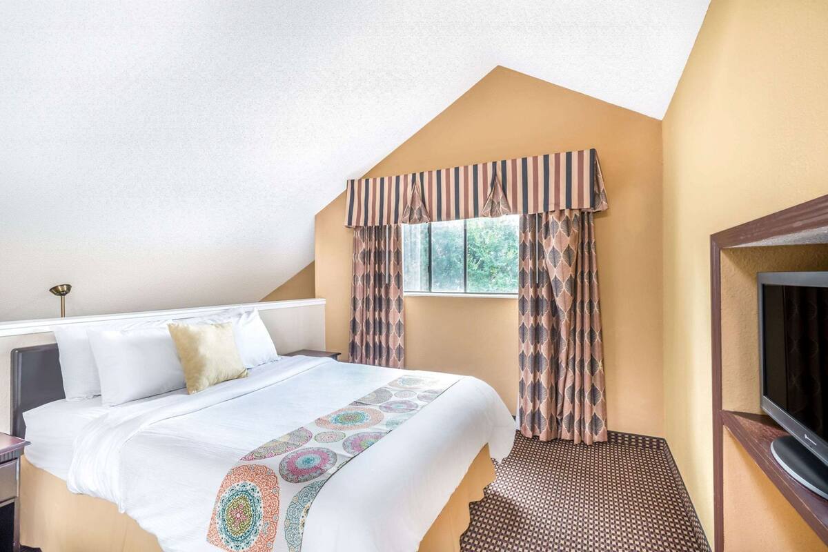 Una habitación de hotel con cama, ventana con cortinas, televisor y una mesita.